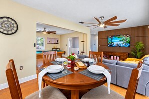 A dining area with a wooden table set for four, a pool table in the background, and a living room with a sofa and a TV.