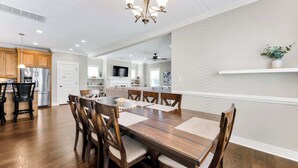 A spacious dining area with a wooden table and chairs, a chandelier, and a kitchen in the background.