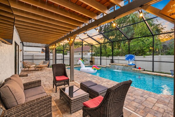 A covered outdoor area with a pool, lounge chairs, and a basketball hoop.