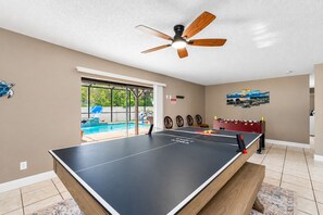 Una habitación con mesa de ping pong, ventilador de techo y vista a una piscina.