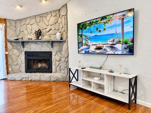 Una sala de estar con chimenea de piedra, un televisor montado que muestra una escena tropical, y piso de madera.