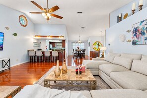 Amplia sala de estar con un sofá grande, una mesa de centro con objetos decorativos, un comedor con sillas y una cocina al fondo.