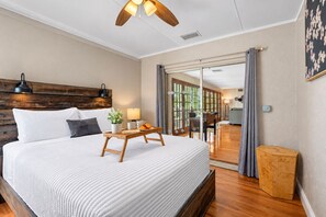 A hotel room with a large bed, a wooden headboard, a bedside table with a lamp, a small plant, and a tray with a cup and a pastry.