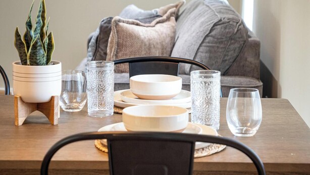 A dining table with white plates, bowls, and clear glasses, set on a wooden table with a potted plant and a decorative vase.