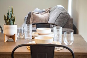 A dining table with white plates, bowls, and clear glasses, set on a wooden table with a potted plant and a decorative vase.