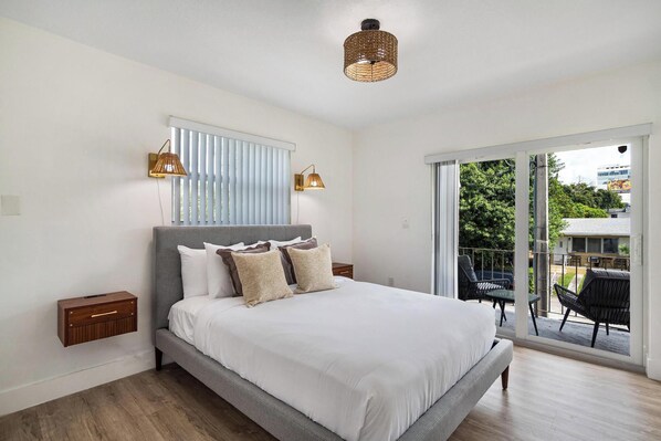 A bedroom with a bed, two bedside tables, a window with blinds, and a view of the outdoors.