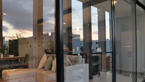 Un salon moderne avec un canapé et des fauteuils, un coin repas avec une table et des chaises, et une vue sur le paysage urbain grâce à de grandes fenêtres.