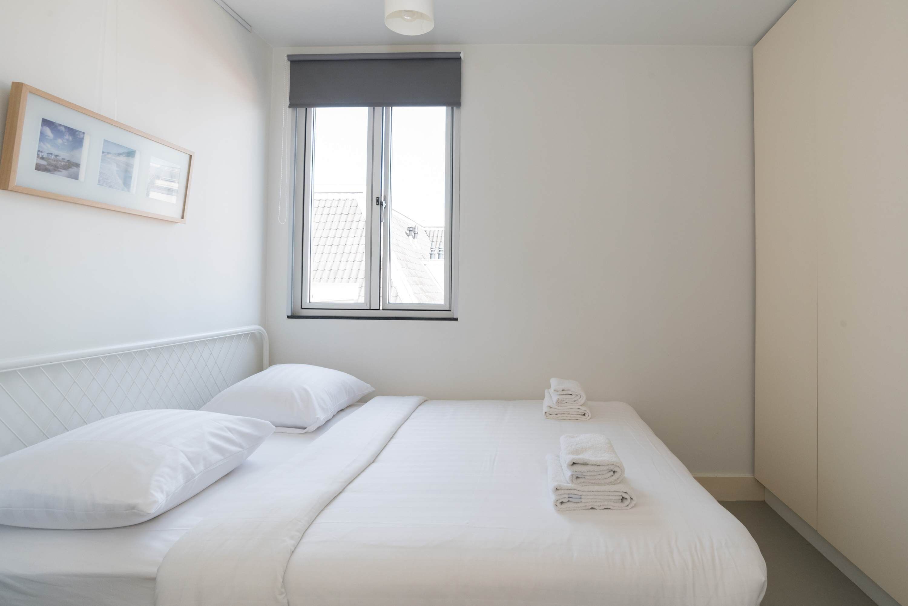 A neatly made bed with white linens and a window with a dark blind.