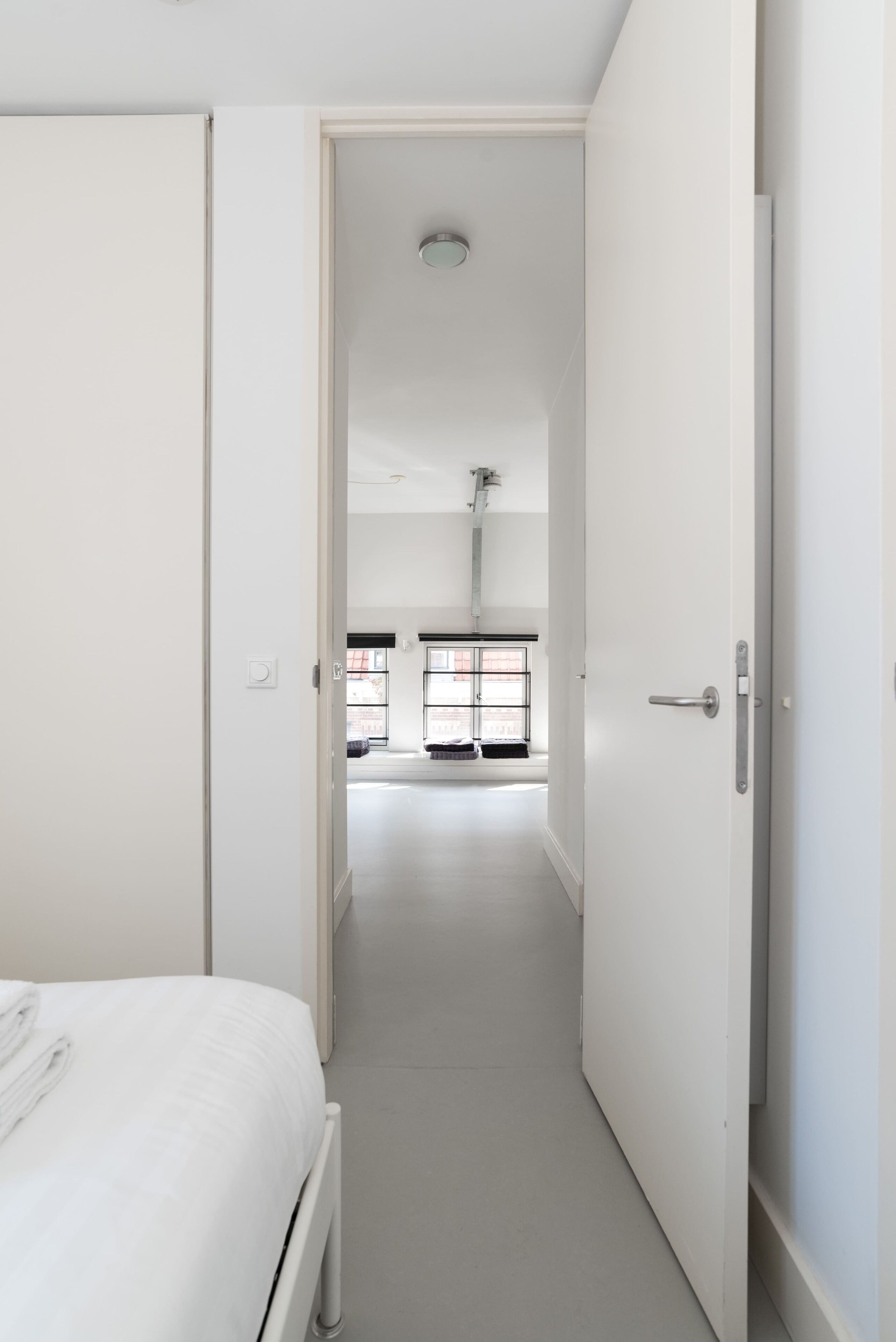 A narrow hallway with a bed on the left and a door leading to a room with a window and a radiator.