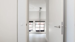 A narrow hallway with a bed on the left and a door leading to a room with a window and a radiator. - Laurier Apartment - in the Jordaan (Amsterdam)