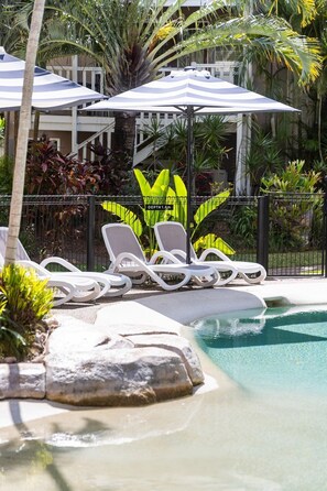 A pool area with lounge chairs and umbrellas, surrounded by tropical plants and a fence.