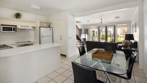 Een moderne keuken met witte kasten, een magnetron en een koelkast. Er is een eethoek met een glazen tafel en stoelen, en uitzicht op een tuin door grote ramen.