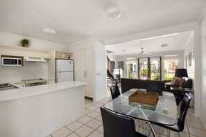 A modern kitchen with white cabinetry, a microwave, and a refrigerator. There is a dining area with a glass table and chairs, and a view of a garden through large windows. - Riverside Rest on Noosa River (Noosa)