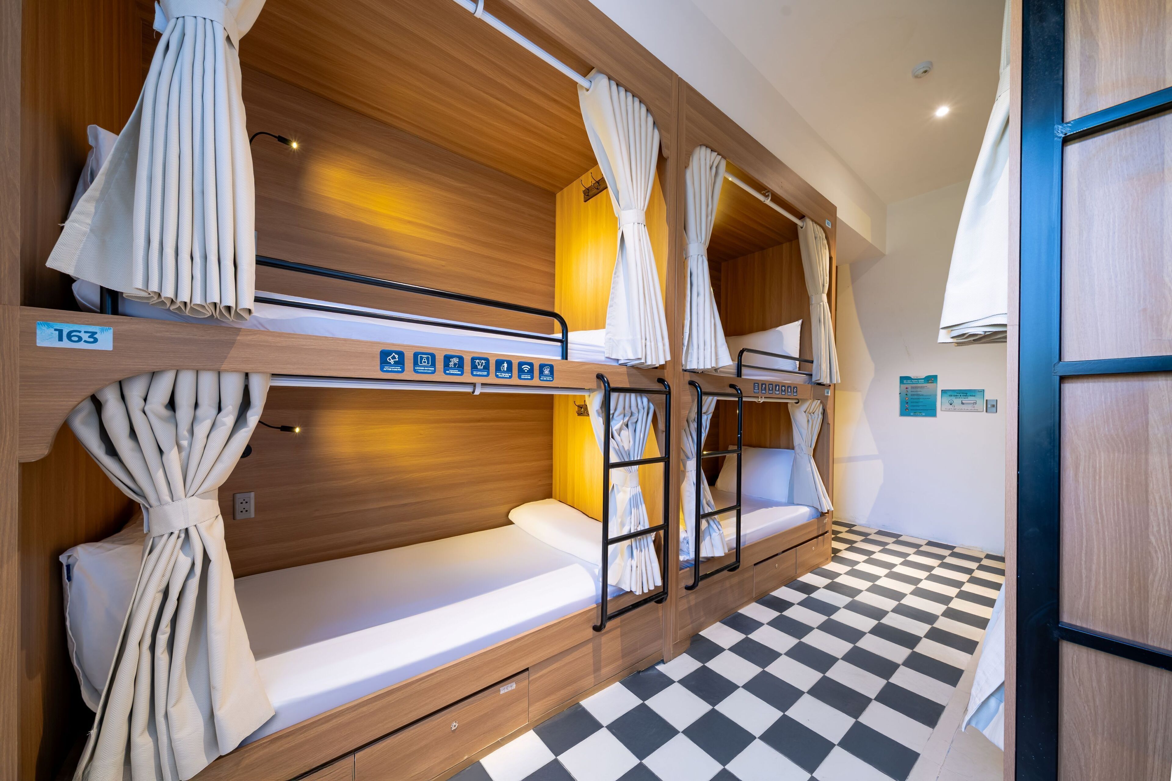 A bunk room with wooden bunk beds, white bedding, and curtains. There are amenities icons on the beds, and a checkered floor.