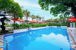 A swimming pool with red umbrellas, surrounded by greenery and palm trees, with a cityscape in the background. - Sujet Beach Hotel &Apartment by Haviland (Da Nang)