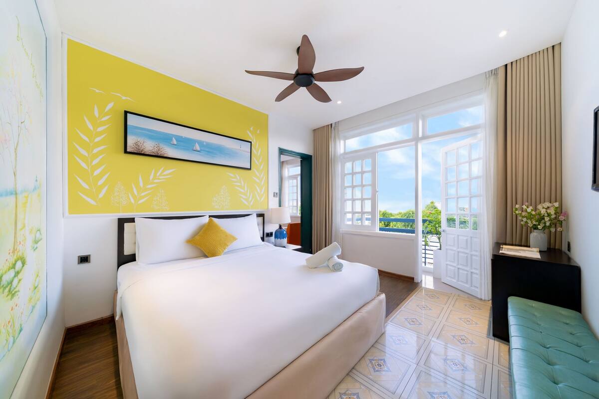 A hotel room with a large bed, a ceiling fan, a window with a view, and a painting on the wall.