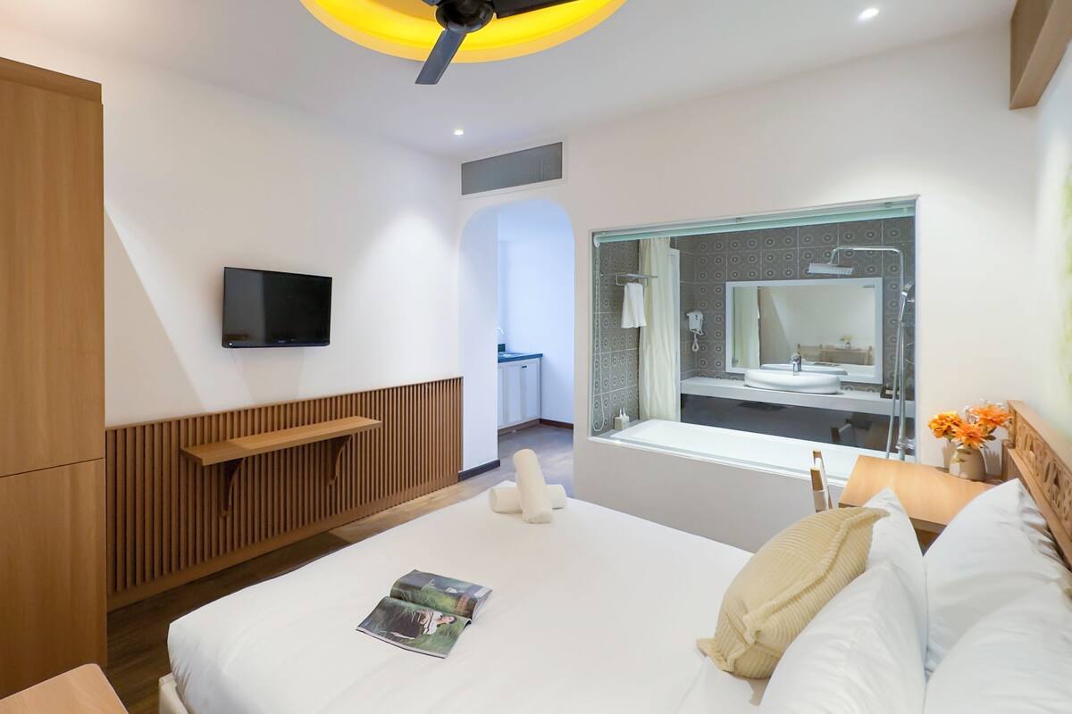 A modern hotel room with a large bed, a TV mounted on the wall, a wooden wardrobe, and a bathroom visible through an open door.