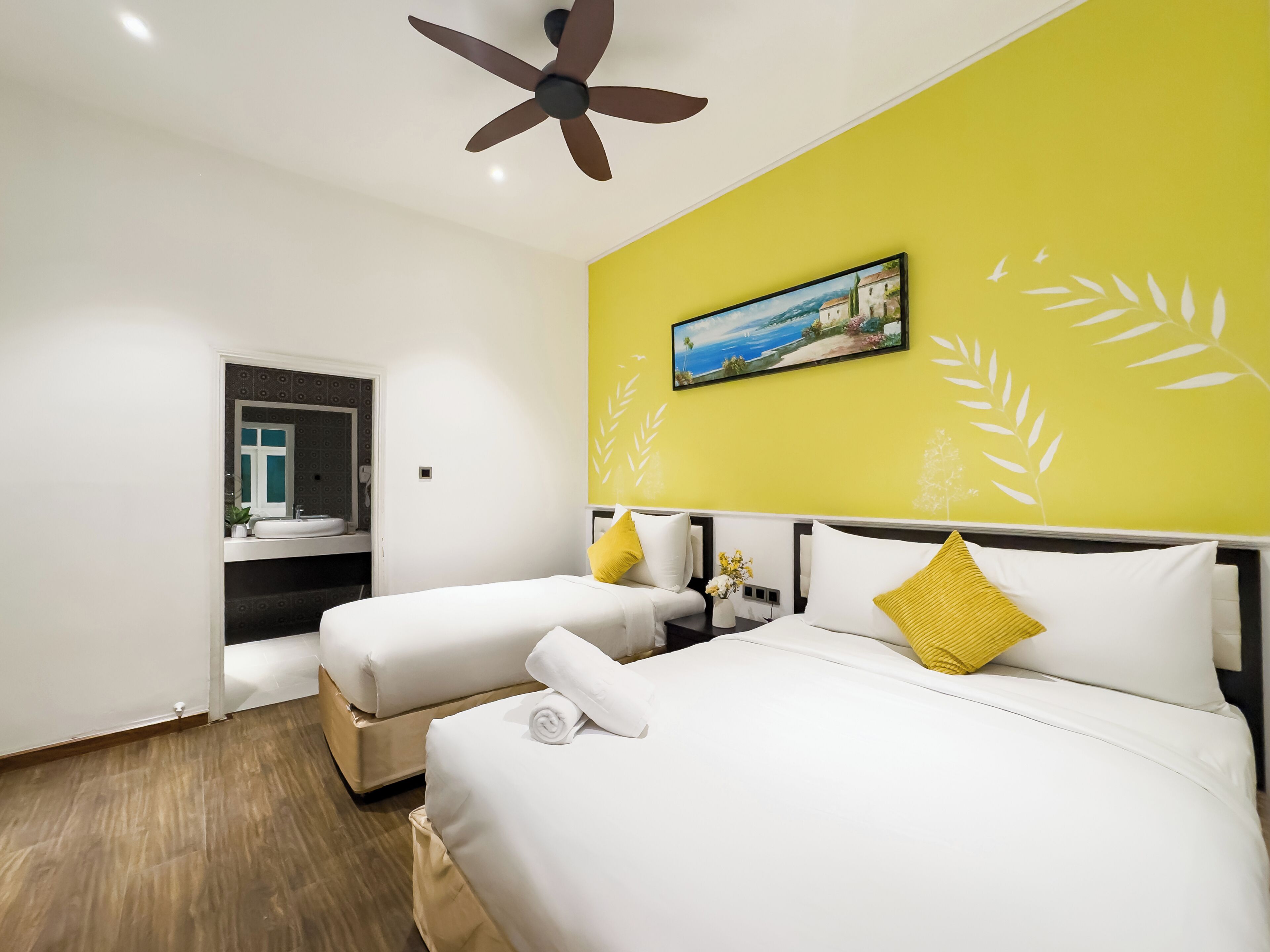 Habitación de hotel con dos camas, un ventilador de techo, un cuadro de una escena costera y una pared amarilla con motivos de hojas.
