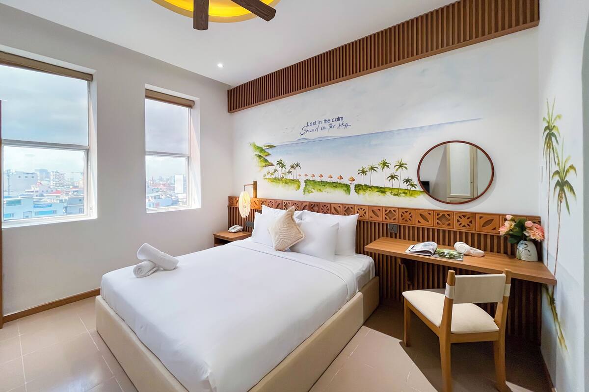 A hotel room with a large bed, a desk, a chair, a ceiling fan, and a city view.
