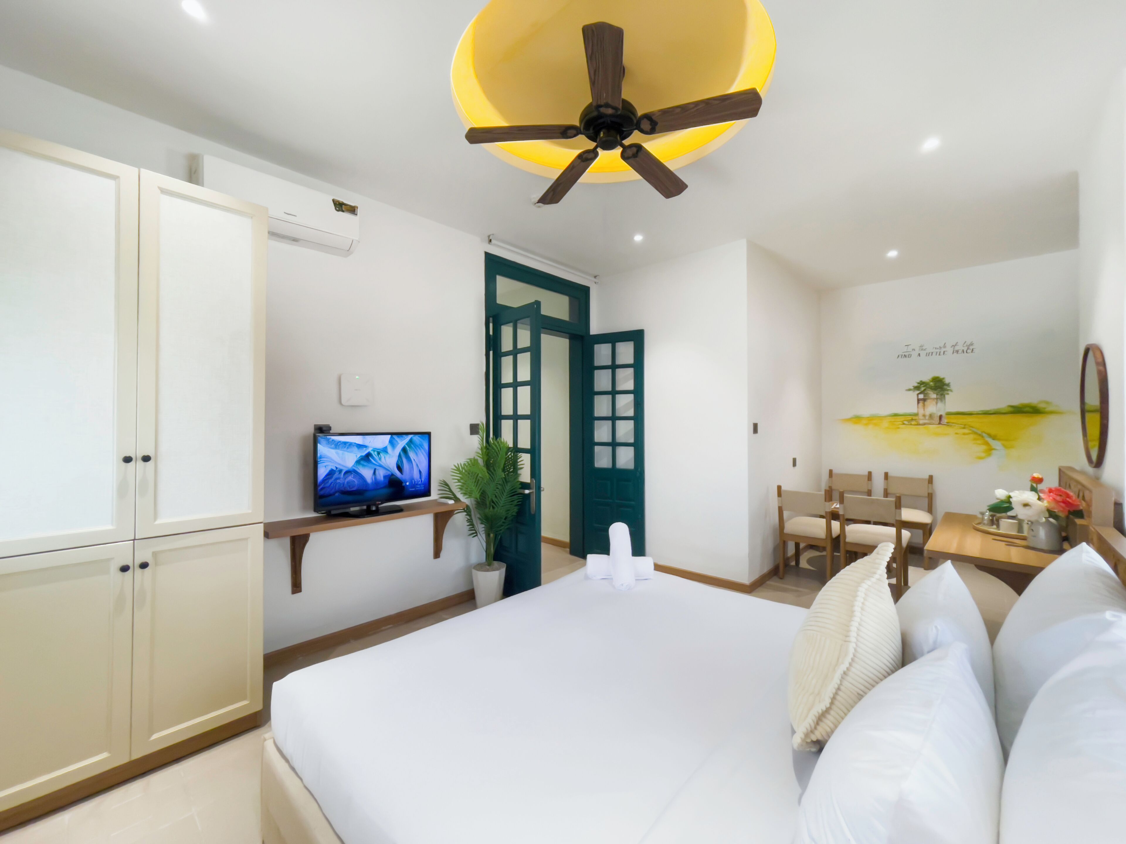 A hotel room with a bed, a TV, a ceiling fan, a desk with a chair, and a wall with a painting.