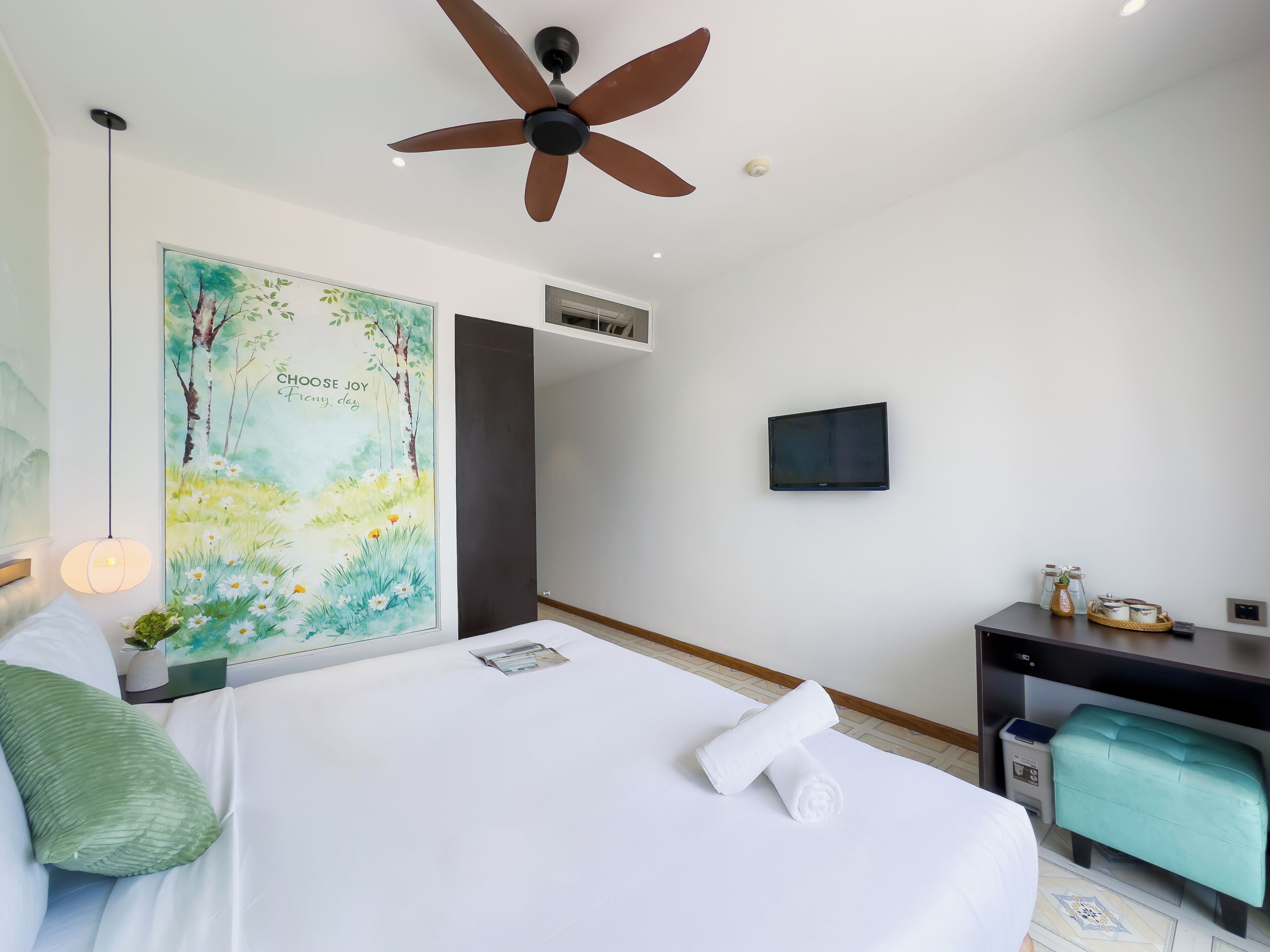 Une chambre d’hôtel avec un lit, un bureau, un ventilateur de plafond, un tableau et un téléviseur fixé au mur.
