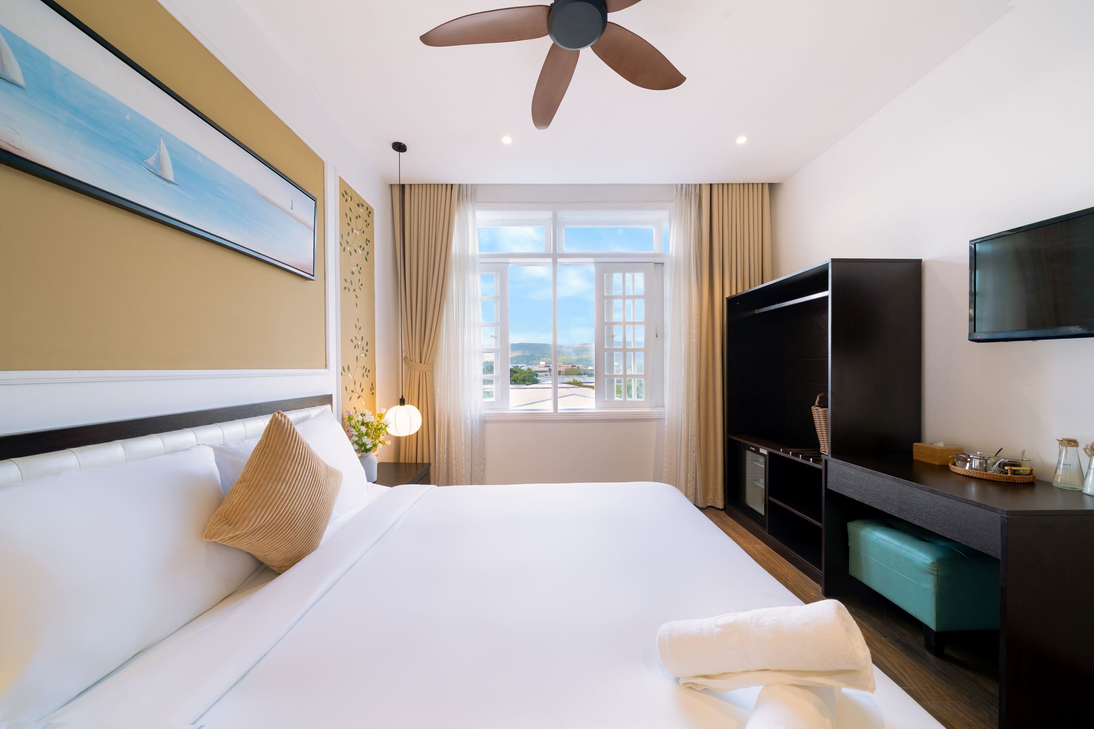 Quarto de hotel com uma cama grande, um ventilador de teto, uma televisão e uma janela com vista para a cidade.