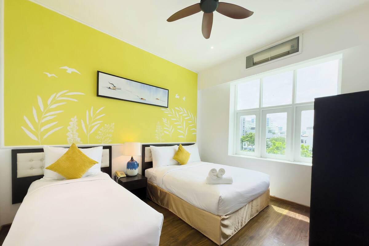A hotel room with two beds, a ceiling fan, a window with a city view, and a painting on the wall.