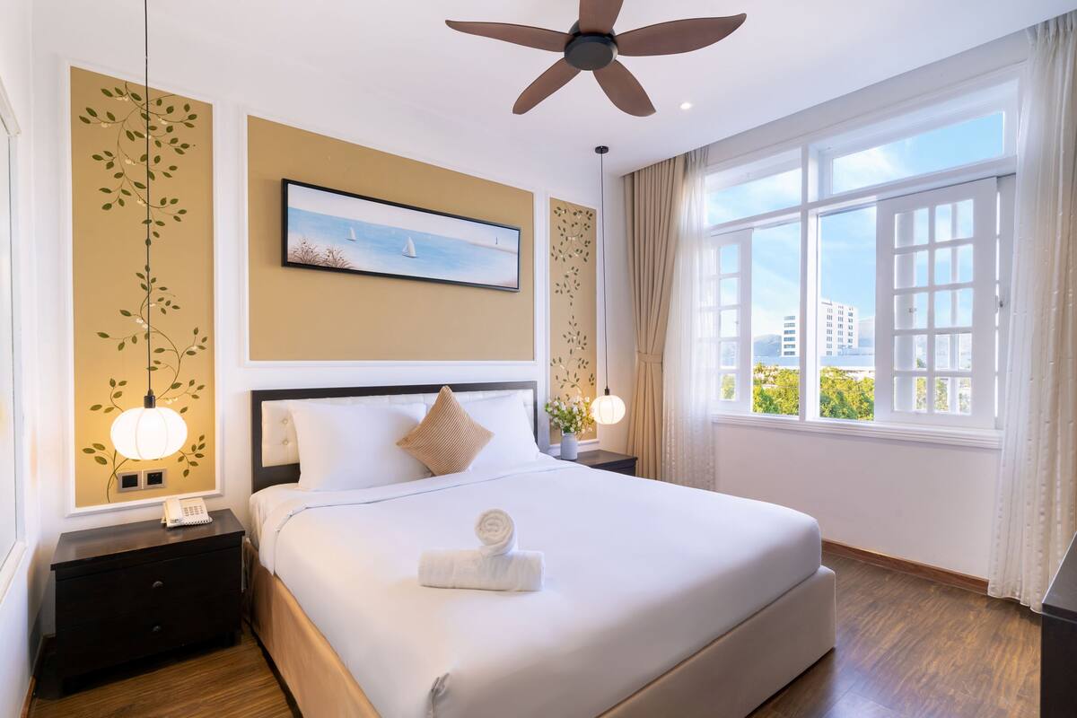 A hotel room with a large bed, a ceiling fan, a window with a city view, and a painting on the wall.