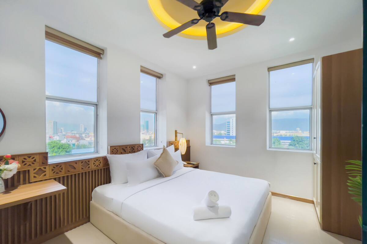 A modern hotel room with a large bed, two windows, a ceiling fan, and a view of the city.