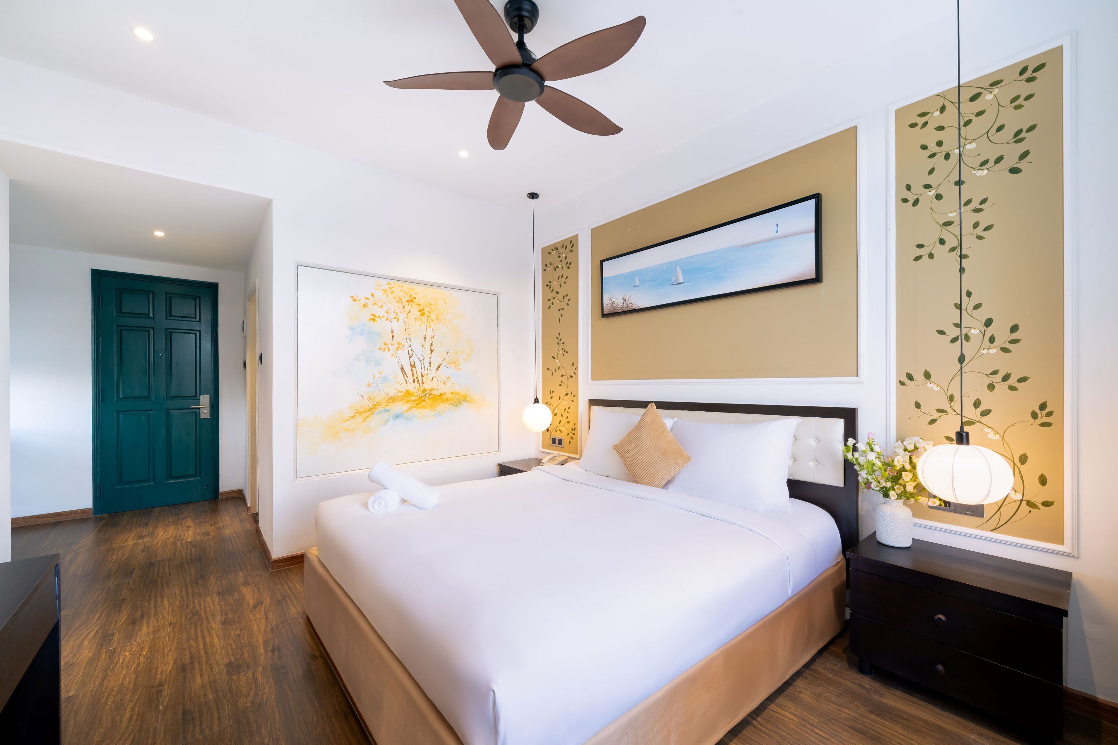 A hotel room with a large bed, a ceiling fan, a painting, a nightstand with a lamp, and a door.