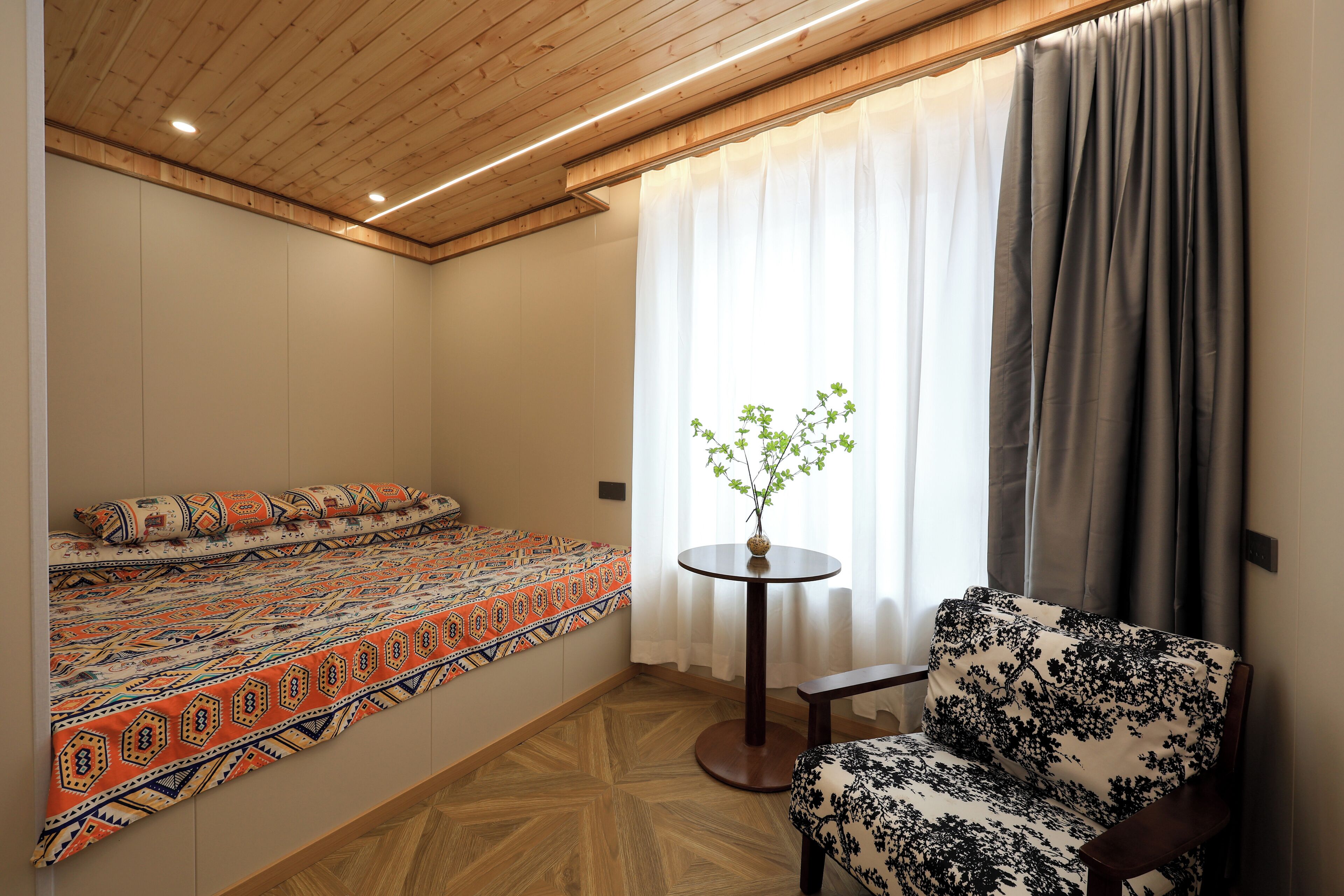 A room with a bed, a chair, a small table with a plant, and a window with curtains.