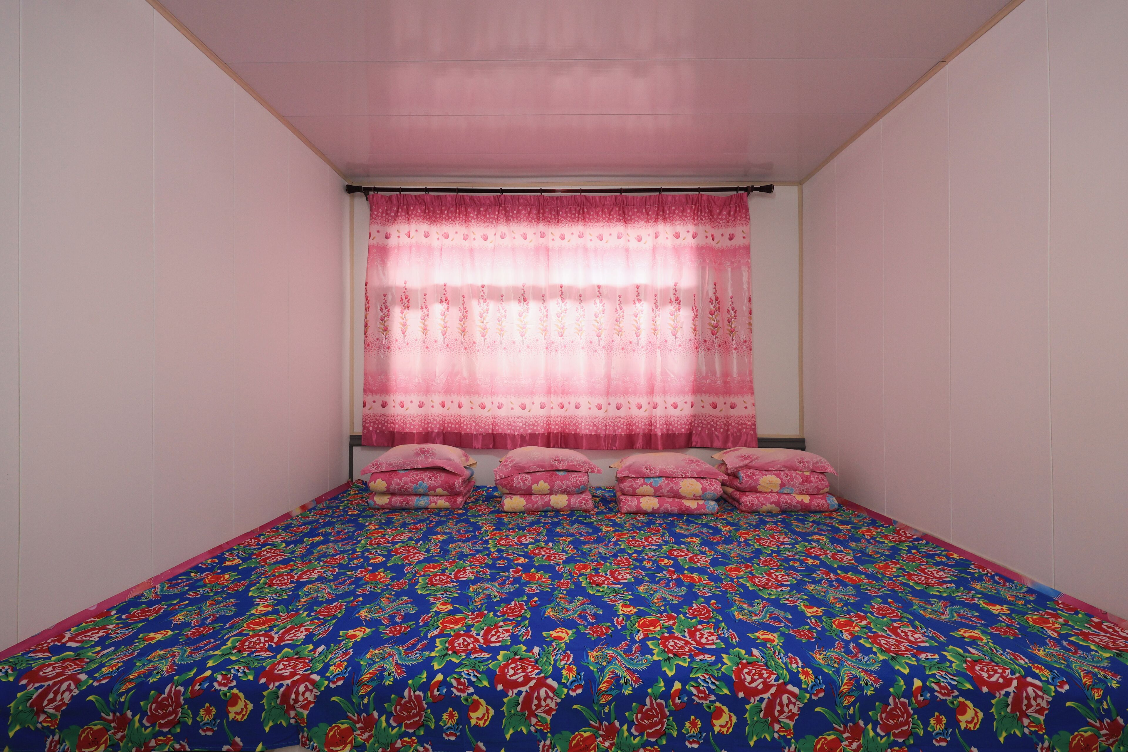 A bedroom with a floral-patterned bedspread, pink curtains, and multiple pillows.