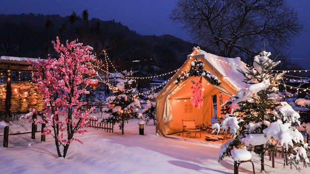 Un espace extérieur accueillant, recouvert de neige, avec une structure ressemblant à une tente, des guirlandes lumineuses et des arbres enneigés.