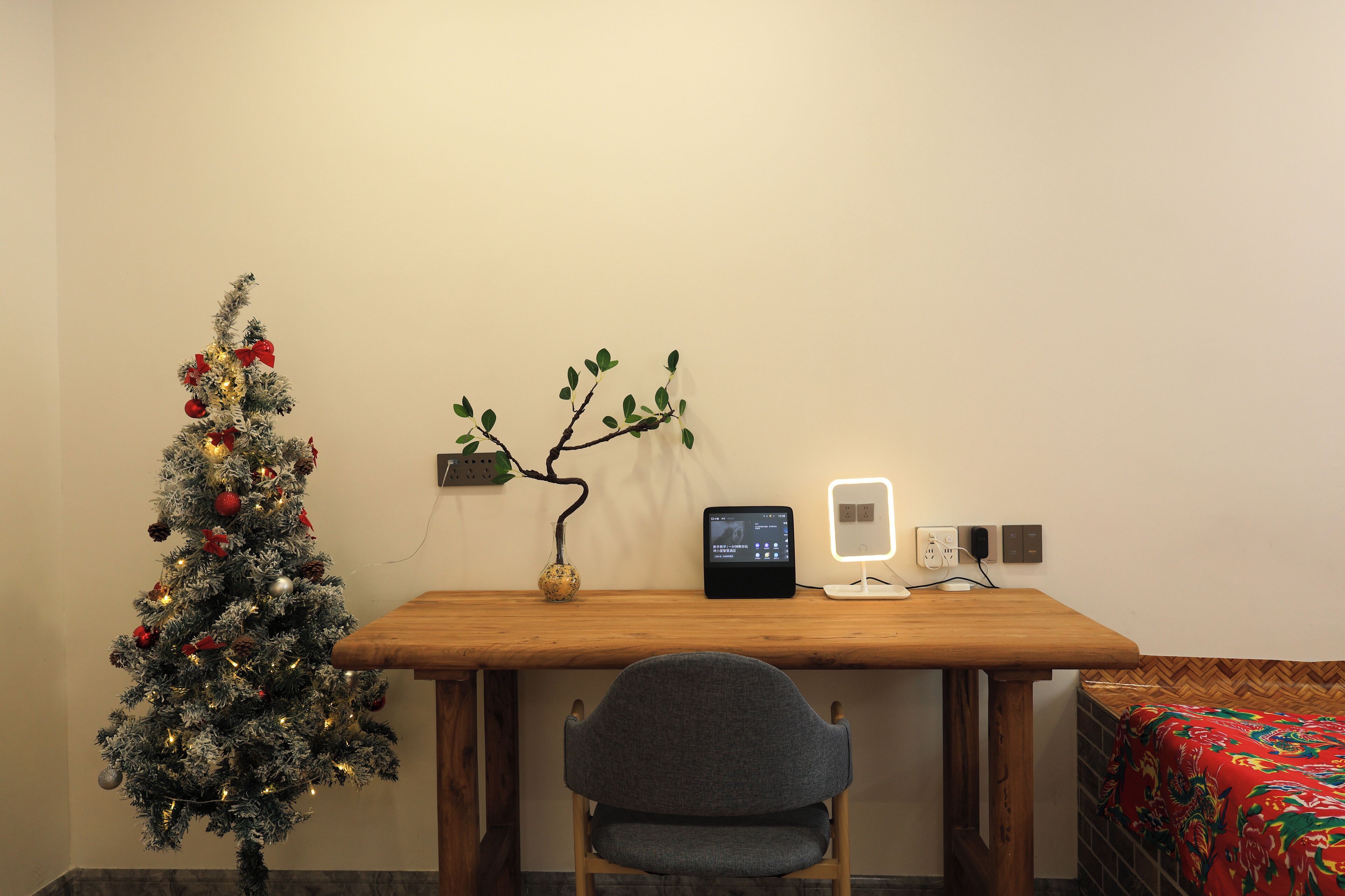 A room with a wooden desk, a small Christmas tree, a potted plant, a tablet, and a bedside table with a lamp.