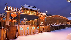 Une porte en bois traditionnelle, dont le toit est recouvert de neige, illuminée par des lumières chaleureuses, et qui mène à un bâtiment sur lequel on peut lire une inscription dans une langue étrangère.