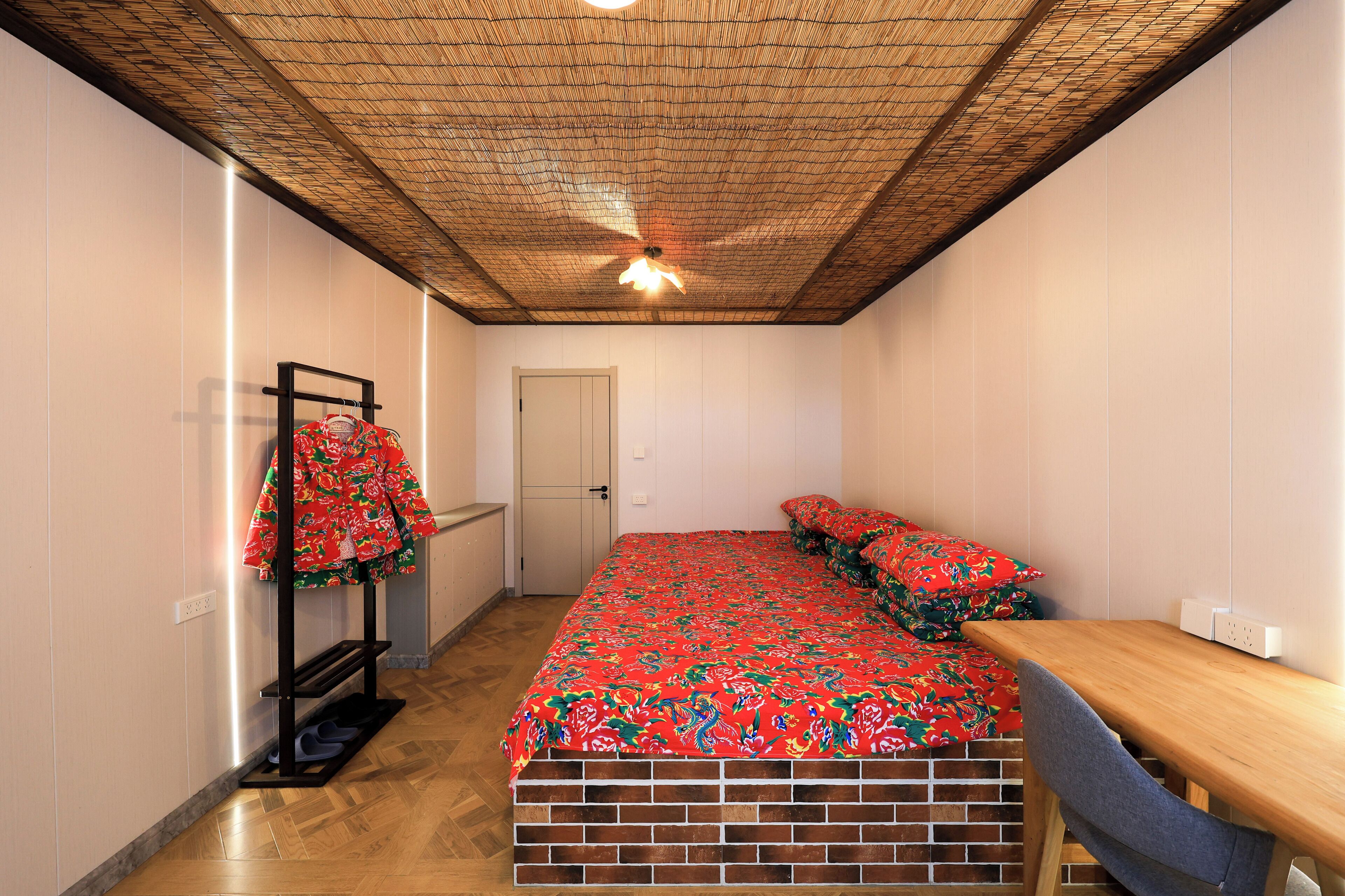 Un dormitorio con una cama, una mesa de madera, una silla, un perchero con un abrigo rojo y una puerta.