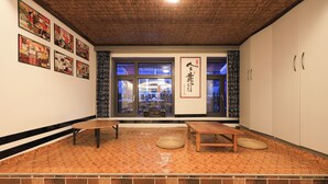 A room with a wooden ceiling, a brick wall, and a large window. There are two wooden tables and a woven mat on the floor.