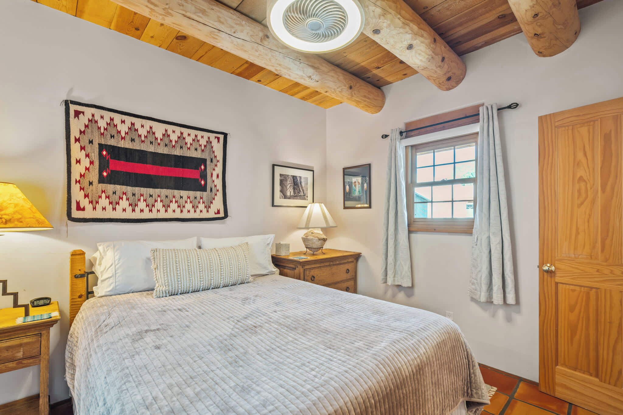 Um quarto com cama, mesa de cabeceira, luminária, janela com cortinas e porta de madeira.