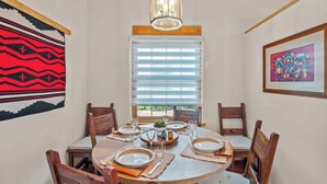 A dining area with a round table set for four, wooden chairs, a chandelier, a window with blinds, and wall art. - Walk to Plaza and Railyard, Fire Pit (Santa Fe)
