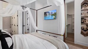 Ein Schlafzimmer mit Baldachinbett, einem an der Wand montierten Fernseher und einem Kleiderschrank mit darin hängender Kleidung.