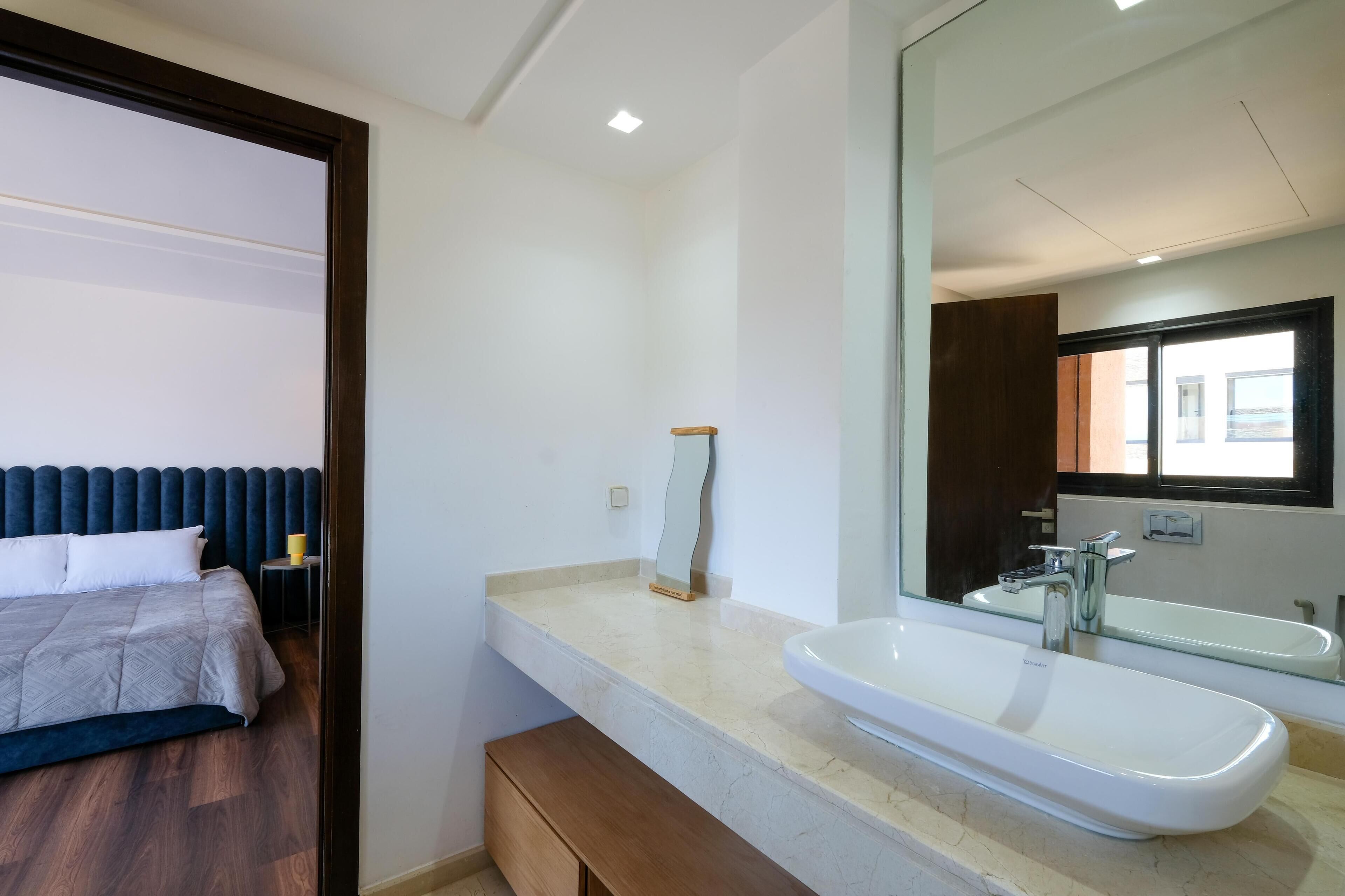 A modern hotel room with a bed, a bathroom with a large mirror, a sink, and a window.