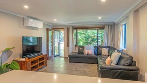 A living room with a flat-screen TV on a wooden stand, a leather sofa, and a large window with curtains.