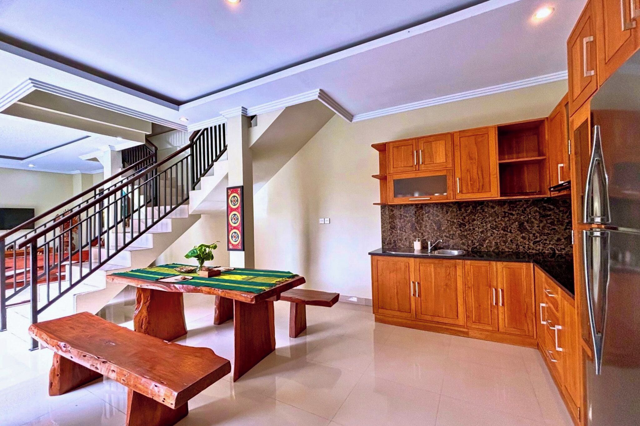 A spacious kitchen with wooden cabinets, a stainless steel refrigerator, a marble countertop, and a dining area with a wooden table and benches.