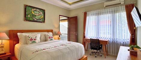 A hotel room with a large bed, a desk, and a chair. There is a window with curtains, an air conditioning unit, and a painting on the wall.