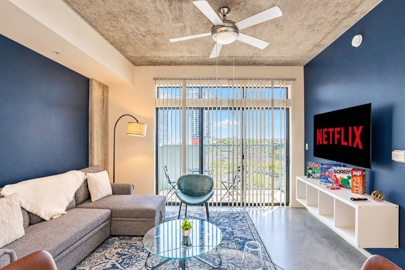 A living room with a sofa, a glass coffee table, a ceiling fan, a TV, and a balcony with a view.