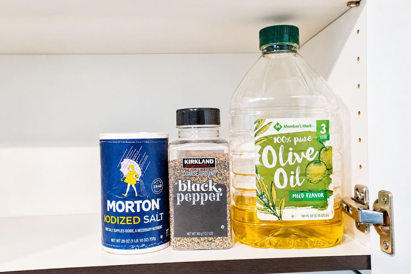 A shelf with Morton iodized salt, Kirkland black pepper, and a bottle of 3-liter pure olive oil.