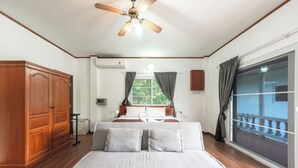 Een slaapkamer met een bed, een houten garderobe, plafondventilator, raam met gordijnen en uitzicht op een balkon.