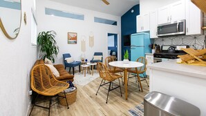 Un espace de vie compact comprenant un coin cuisine, un coin repas avec des chaises en osier, un canapé et un ventilateur de plafond.