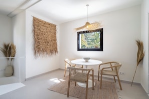 A minimalist dining area with a round table, two chairs, a woven wall hanging, and a framed picture of a plant.