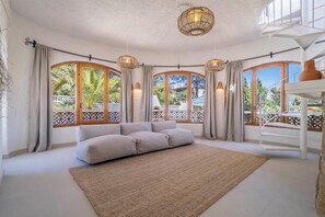 A spacious room with a large sofa, a woven rug, and a view of greenery through large windows.